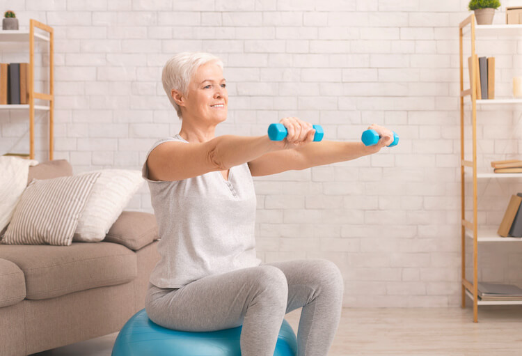 Ernährung für Senioren 1 Ernährung für Senioren. Eine Senioren sitzt auf einem Gymnastikball in ihrem Wohnzimmer. Sie streckt beide Arme nach vorne und hält in jeder Hand eine kleine, blaue Hantel.