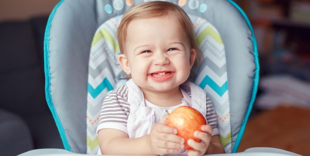 Kleinkind sitzt im Hochstuhl und isst einen Apfel – ausgewogene Ernährung für Kleinkinder im Familienalltag.